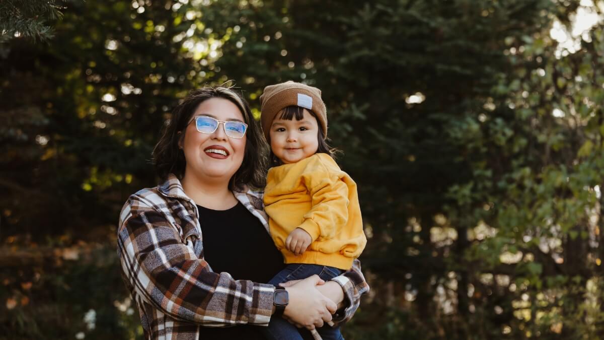 Indigenous woman holding an Indigenous toddler girls standing in front of trees outside